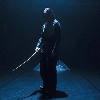 Szene aus Near Misses. Dunkle Bühne, einzelne Figur im Lichtkegel mit langem, gebogenem Objekt in der Hand.