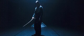 Szene aus Near Misses. Dunkle Bühne, einzelne Figur im Lichtkegel mit langem, gebogenem Objekt in der Hand.