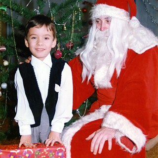 Ein Kind steht vor einem geschmückten Weihnachtsbaum neben einer Person in einem roten Kostüm mit weißem Bart und Mütze.