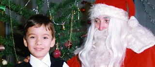 Ein Kind steht vor einem geschmückten Weihnachtsbaum neben einer Person in einem roten Kostüm mit weißem Bart und Mütze.