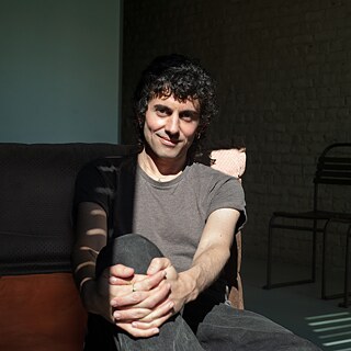 El artista Alberto Cortés está sentado en una silla en una habitación, iluminado por el sol. Mira a la cámara.