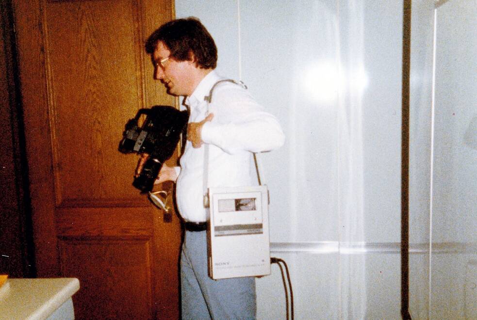 Eine Person hält eine große, ältere Videokamera in den Händen und trägt einen tragbaren Videorekorder über der Schulter. Die Geräte wirken sperrig und stammen aus einer früheren Generation der Aufnahmetechnik. Die Person steht in einem Innenraum, der Hintergrund ist unscharf. Die Szene vermittelt den Eindruck privater, dokumentarischer Aufnahmen aus der Vergangenheit.