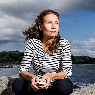 Die Fotografin sitzt auf Steinen am Meer und blickt nach rechts in die Ferne.