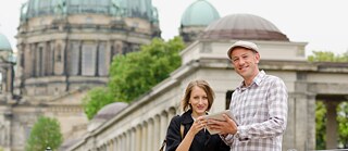 Zwei Personen nutzen gemeinsam ein Tablet vor historischer Berliner Architektur mit Kuppeln und Säulen.