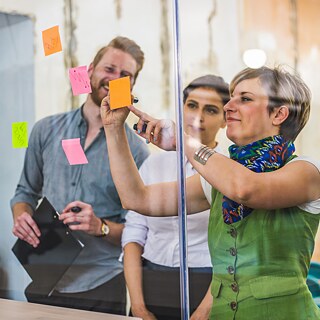	Drei Teamkollegen arbeiten kreativ mit Post-its an einer Glaspinnwand. Sie sind sehr konzentriert und haben Spaß.