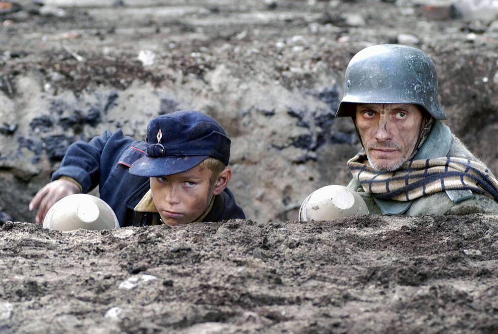 Das Standbild aus „Der Untergang“ (2004) zeigt einen sehr jungen kindlichen Volksturm Soldaten mit einer Panzerfaust gemeinsam mit einem älteren hageren Wehrmachtssoldaten in einem Schützengraben. Die Darstellung der letzten Tage des dunkelsten Kapitels der deutschen Geschichte in einem abendfüllenden Spielfilm: dem Produzenten Bernd Eichinger und Regisseur Oliver Hirschbiegel gelangen mit dem monumentalen Epos einer der bedeutendsten und meistdiskutierten Kriegsfilme seit „Das Boot“. Mit einem herausragenden Ensemble inszeniert das Werk die letzten zwölf Tage der nationalsozialistischen Herrschaft und ihren endgültigen Zusammenbruch.