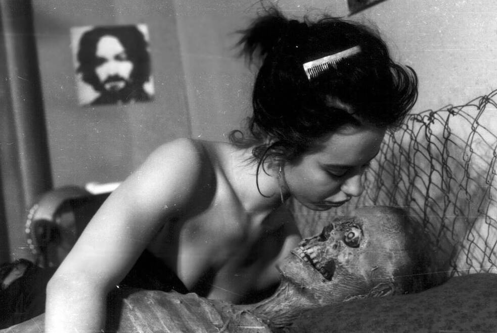 Standbild aus "Nekromantik" (1988) zeigt eine junge Frau, die sich über einen zurückgelehnten, verwesenden Körper beugt, um ihn zu küssen. Der Independent Horrorfilm von Jörg Buttgereit erlangte durch seine extremen Themen und gewagt experimentelle Form internationale Aufmerksamkeit. Der Film gilt als Kultklassiker des Undergroundkinos und als eines der umstrittensten Werke des Horrorgenres.