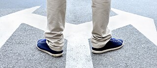 Untere Hälfte einer stehenden Person auf einer Straße, die zwischen zwei entgegengesetzten Richtungs­pfeilen positioniert ist. Die Person trägt helle Hosen und dunkle Schuhe, und die Pfeile auf dem Asphalt symbolisieren zwei mögliche Wege