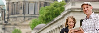 Zwei Personen nutzen gemeinsam ein Tablet vor historischer Berliner Architektur mit Kuppeln und Säulen.