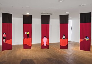 Installationsansicht mit fünf von der Decke hängenden Textilbannern in Schwarz und Rot; jedes Banner zeigt eine figurative textile Darstellung einer einzelnen Person auf einer roten Fläche, präsentiert in einem modernen Ausstellungsraum mit Holzfußboden und weißem Hintergrund.