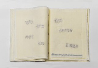 Offenes textiles Buch aus hellen Stoffseiten, auf denen der Satz „We are not on the same page“ sowie eine kleine zusätzliche Stickschrift eingearbeitet ist; kunsthandwerkliches Textilobjekt in Beige  und Cremetönen.