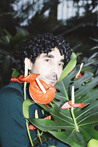 Person mit dunklen, lockigen Haaren posiert zwischen großen tropischen Blättern und leuchtend roten Blumen und blickt mit starkem Blitzlicht in die Kamera.