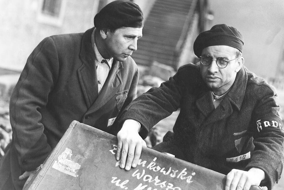 Zwei Männer in dunklen Mänteln und Mützen stehen eng beieinander und wirken in ein ernstes Gespräch vertieft. Einer trägt eine Armbinde mit der Aufschrift „JUD“. Beide halten oder stützen einen Koffer, auf dem handgeschriebene Worte zu erkennen sind. Die Szene ist in Schwarz‑Weiß aufgenommen und zeigt eine städtische Umgebung mit Treppe und Gebäudewand im Hintergrund. Die Stimmung ist angespannt und dokumentarisch.