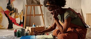 A woman sits in the foreground of an art studio, focused on painting a canvas.
