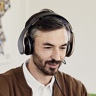 Eine Lehrkraft im Sprachkurs am Goethe-Institut. Eine männliche Lehrkraft gibt online einen Deutschkurs. Er trägt Kopfhörer, spricht mit den Teilnehmern und arbeitet gleichzeitig am Laptop.