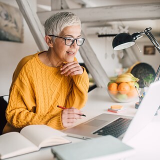 Eine ältere Frau arbeitet im Homeoffice an ihrem Laptop. Konzentriert verfolgt sie ihre Aufgaben und hält wichtige Gedanken in Notizen fest. 