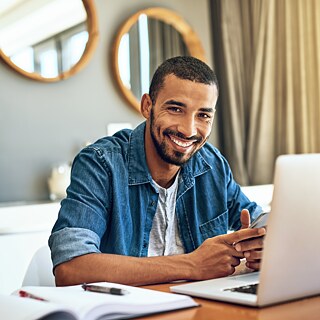 Ein Mann sitzt zu Hause vor einem Laptop und arbeitet. Er lächelt selbstbewusst in die Kamera.