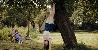 Ein Garten, in dem link im Hintergrund jemand im Gartenstuhl sitzt und im Vordergrund ein junges Mädchen einen Handstand gegen einen Baum macht.