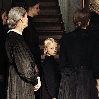 Eine Gruppe von Menschen in altmodischer Kleidung ist von der Seite zu sehen, wie sie nach rechts blicken. In der Mitte schaut ein kleines Mädchen, auf das das Licht fällt, den Betrachter an.