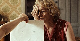 A woman with blonde hair and glasses is covering her right eye and looking with her other eye at a sheet of paper bearing two symbols, which is being held up by a hand reaching into the picture.