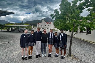 Sechs Schüler*innen in Uniform und eine erwachsene Person in der Mitte Stehen auf einem mit Kies bedeckten Platz. Neben ihnen ein belaubter Baum, hinter ihnen ein Gebäude, ein überdachter Parkplatz; im Hintergrund Berge. Der Himmel ist wolkenverhangen, stahlgrau.