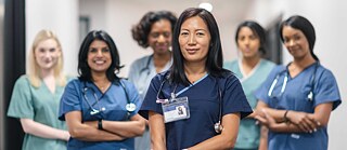Ein Team von Krankenschwestern und Ärzten steht gemeinsam im Korridor des Krankenhauses. Die Gruppe schaut selbstbewusst in die Kamera, sie lachen. 