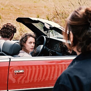 Filmstill fra "Miroirs No. 3". Et par sidder i en åben cabriolet, en kvinde kigger over mod dem.