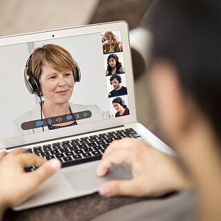Blick über die Schulter eines Kursteilnehmers auf das Laptop mit der Lehrkraft und weiteren Kursteilnehmern auf dem Screen.