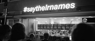 Black and white photo showing a row of people seen from behind who are looking at a row of photographic portraits propped up against a shop window. Above the window a illuminated neon sign says #saytheirnames 