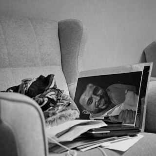 Black and white photo showing a row of people seen from behind who are looking at a row of photographic portraits propped up against a shop window. Above the window a illuminated neon sign says #saytheirnames 