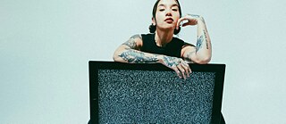 Person sitzt hinter einem Fernseher mit statischem Rauschen; Arme locker über den Bildschirm gelegt.