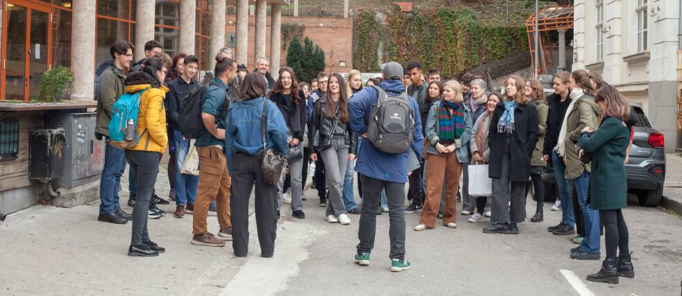 Eine große Gruppesteht in einer Seitenstraße, rechts und links befinden sich Gebäude. Ein Tour-Guide der Graffiti-Tour steht mit dem Rücken zur Gruppe, die Teilnehmenden hören ihm zu. 