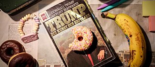 Kniha Die Kunst des Deals na stole a na přebalu leží donut. Vedle knihy vlevo leží další donuty a  vpravo leží banán.