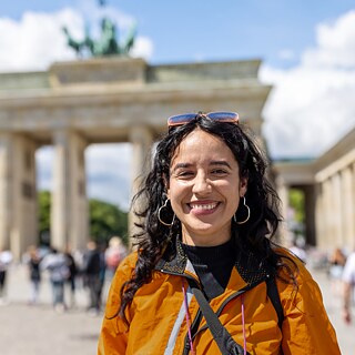Eine Frau lächelt strahlend in die Kamera. In ihrem orangefarbenen Outfit strahlt sie Stolz und Freude aus. Sie steht am Pariser Platz in Berlin, nahe dem Brandenburger Tor. Die beeindruckende Kulisse unterstreicht die besondere Stimmung des Moments.