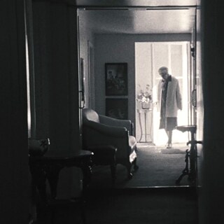 View down a hall way at the end of which we see the silhouette of an old woman against the light room behind her