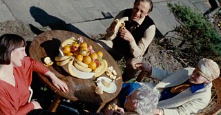 Four people, older and younger sit around a table with a big bowl of fruit on it