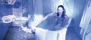 A white woman with long black hair is sitting in a bathtub in a stage-like setting bathed in blue-grey light.