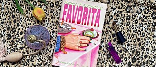 The book “Favorita” lies on a fur rug alongside a yellow tulip, a purple perfume bottle, a lipstick and a purple lighter