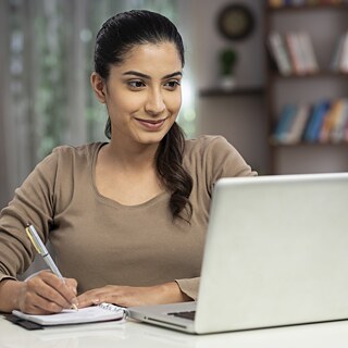 Eine junge Frau sitzt zu Hause am Laptop und lernt. Sie blickt zum Bildschirm und macht sich Notizen.