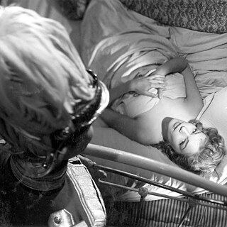 Eine Frau im Bett schaut zu einem Uniformierten auf.