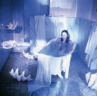 A white woman with long black hair is sitting in a bathtub in a stage-like setting bathed in blue-grey light.