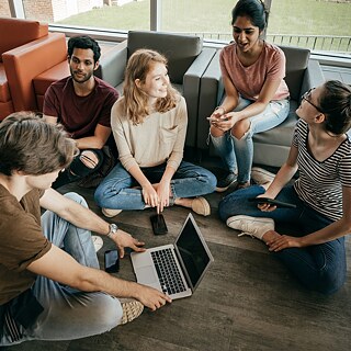Mehrere Studenten sitzen in einem Aufenthaltsraum auf dem Boden und auf Sesseln und unterhalten sich.