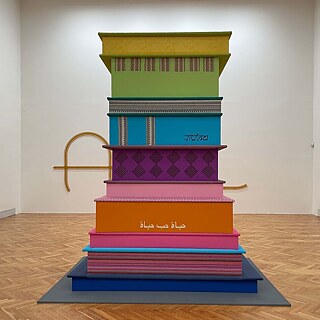 Ein buntes Kunstwerk in einem Ausstellraum. Es sind mehrere niedrige Blöcke, die zusammen einen Turm bilden.
