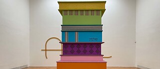 Ein buntes Kunstwerk in einem Ausstellraum. Es sind mehrere niedrige Blöcke, die zusammen einen Turm bilden.