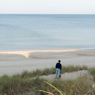 Szene aus dem Film „Kruso“: Person steht in Dünen und blickt auf ruhiges Meer und Sandstrand.
