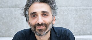 Portrait of a smiling man with gray-black hair and a beard in front of a light gray stone staircase