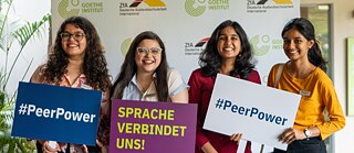 Group of PASCH alumni standing in front of a Goethe‑Institut and ZfA logo wall, holding signs reading “#PeerPower” and “Language connects us!”.