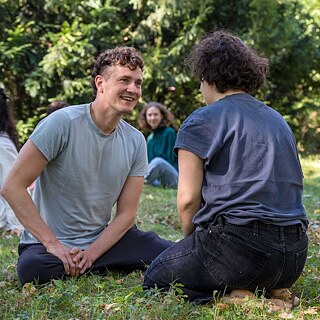 Eine Gruppe von jungen Menschen sitzt beim schönen Wetter im Gras. Im Vordergrund ist ein junger Mann, der gegenüber einer Frau sitzt. Ihm kann man ins Gesicht schauen, die sieht man nur von Hinten.