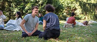 Junge Personen sitzen bei schönem Wetter im Gras. Im Vordergrund ist ein Mann der gegenüber einer Frau sitzt. Ihm sieht man ins Gesicht, sie sieht man von Hinten. 
