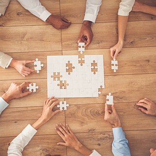 Aufnahme von oben auf einen Tisch, an dem Leute ein Puzzle zusammen setzen. Die Puzzleteile sind weiß.